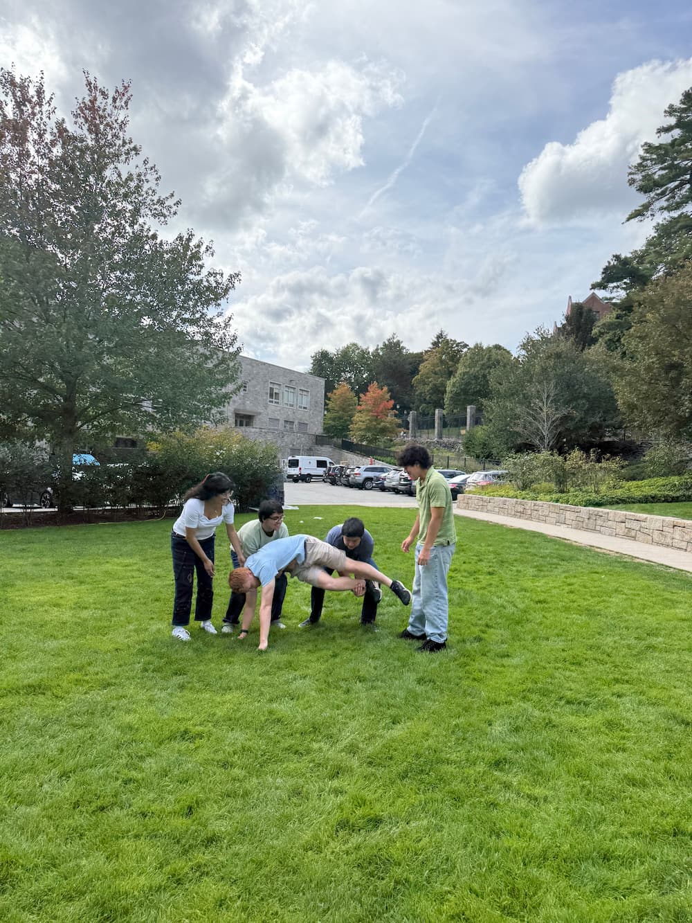 5 students on grass attemting to lift one of them up as a group.