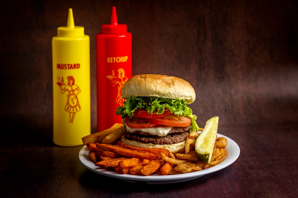 Hamburger with french fries, sweet potato fries, and a pickle in front of ketchup and mustard bottles.
