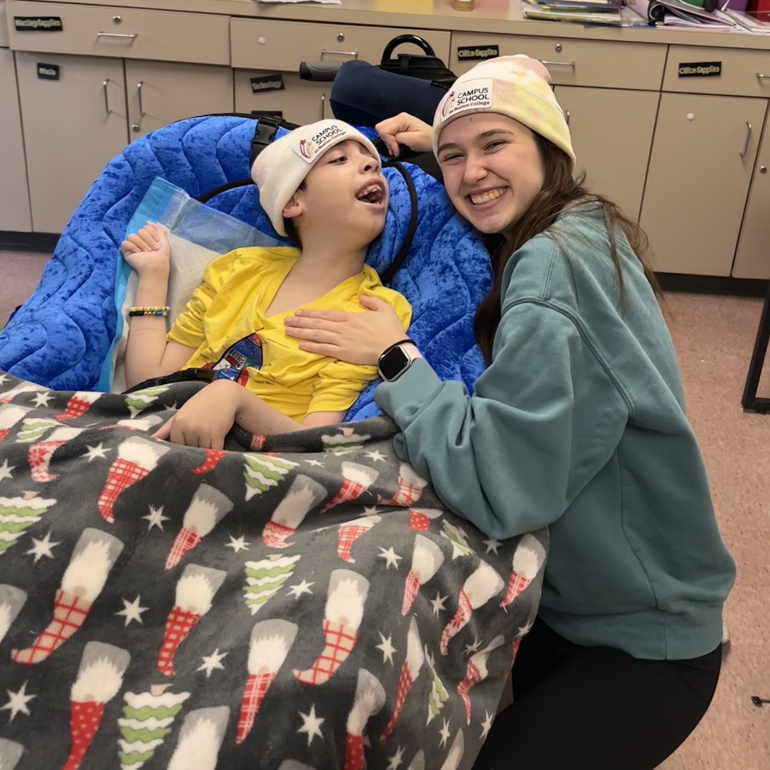 A young boy sitting in a peapod chair next to his teacher in matching Campus School hats