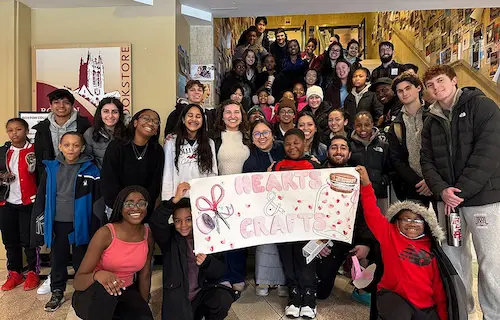 Boston College students and local youth smiling together and holding a big sign during a mentoring program event.