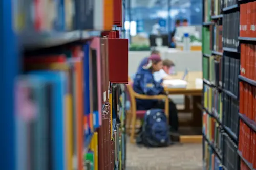 Students studying at a desk in the Boston College library, viewed between rows of bookshelves.