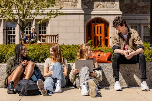 Boston College students sitting outside on campus, talking and working together on a laptop.