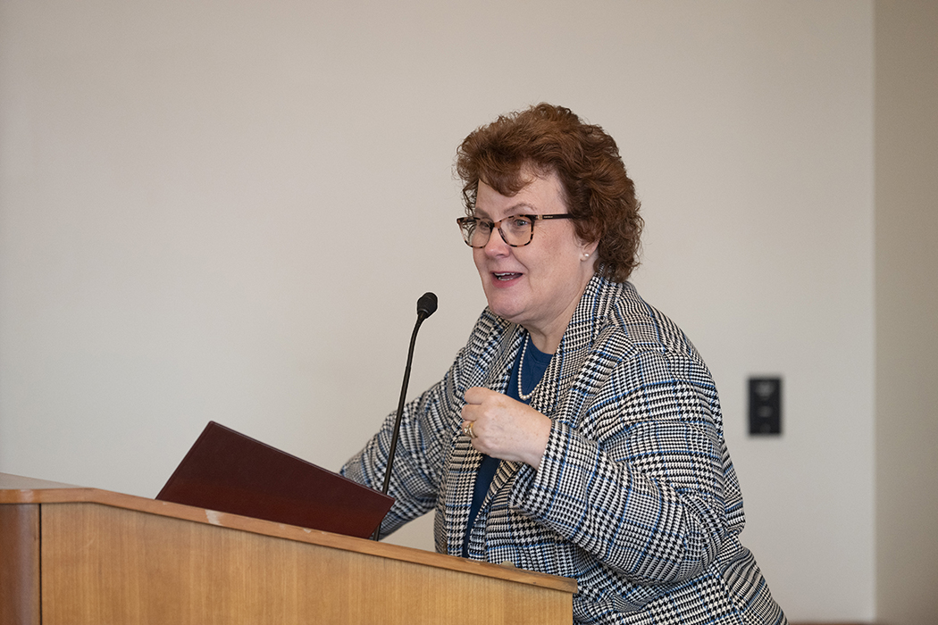 Sue Coleman, in a checkered blazer, speaks behind a podium.