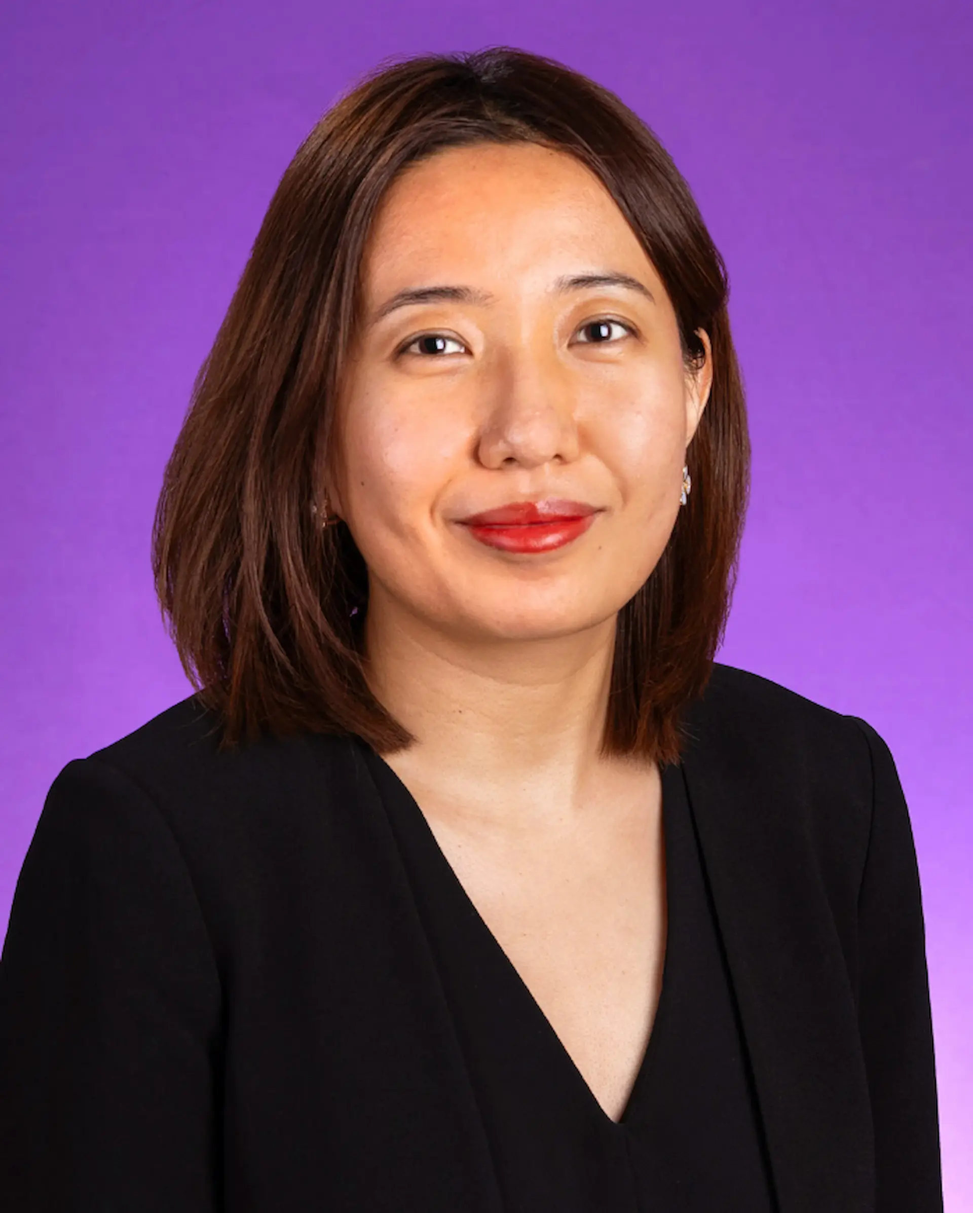 Headshot of a woman in a black top with short brown hair posing in front of a purple background.