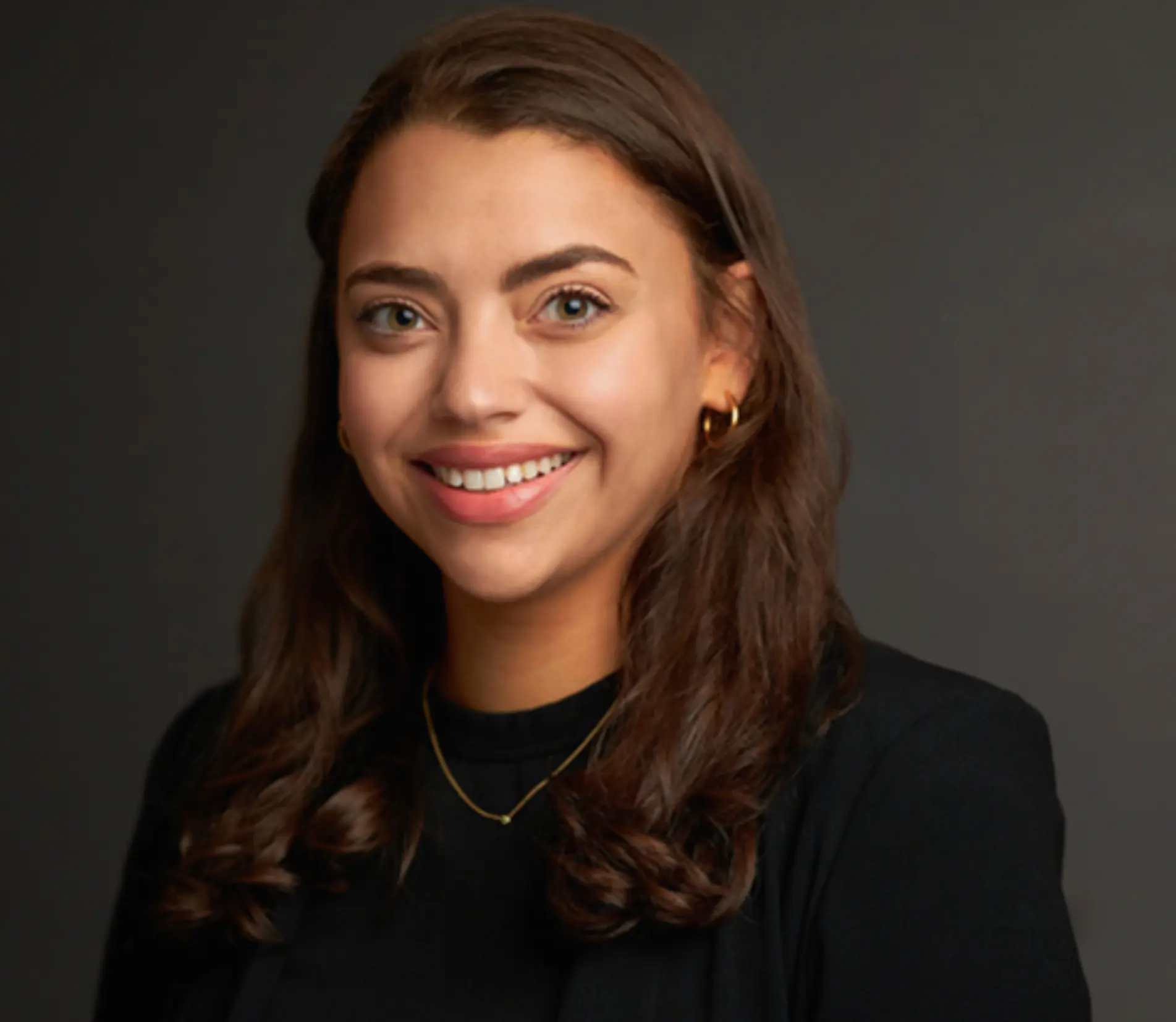 Headshot of a woman with brown wavy hair in a black top smiling in front of a neutral background.