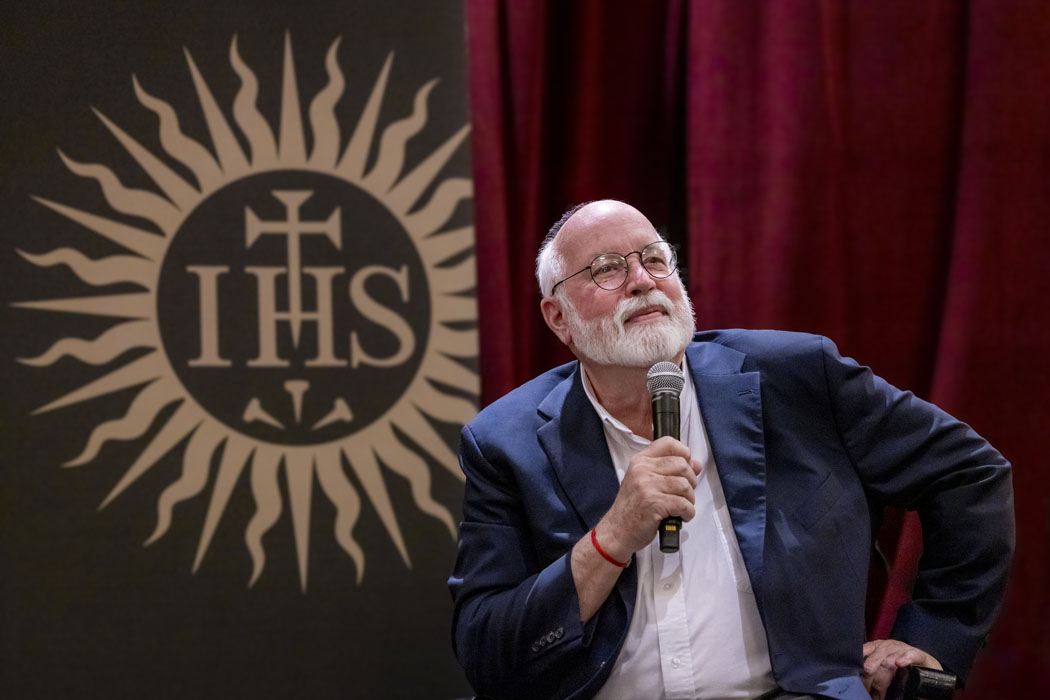 A man with a microphone speaking in front of the Society of Jesus seal
